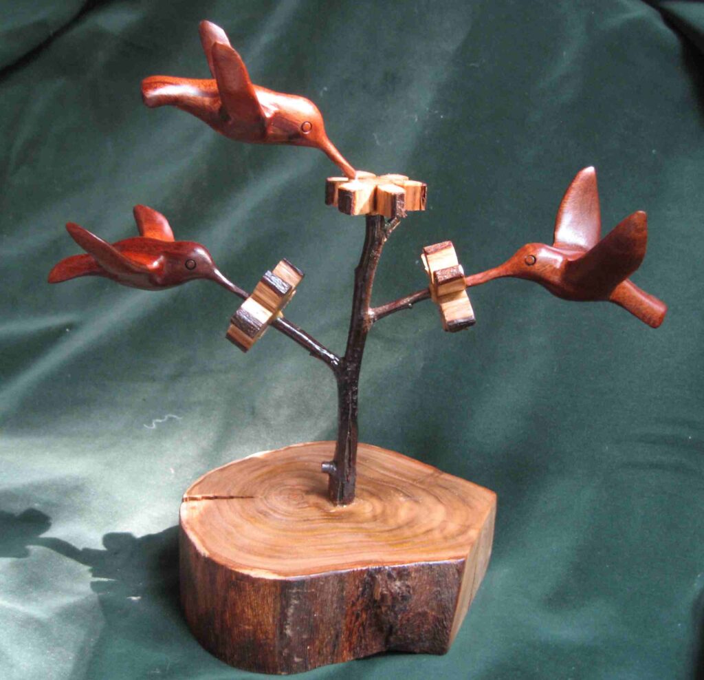 three stylized wood carvings of hummingbirds attached to a display of wooden flowers by gary carver of carverscarvings